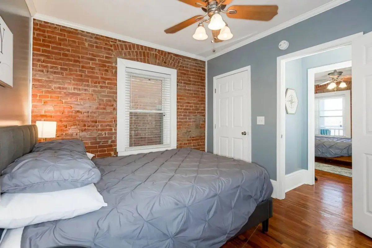 A calming bedroom featuring a bed with gray bedding, an exposed brick wall, ceiling fan, and a large window letting in natural light. The room is simple yet comfortable, perfect for relaxation or reading.
