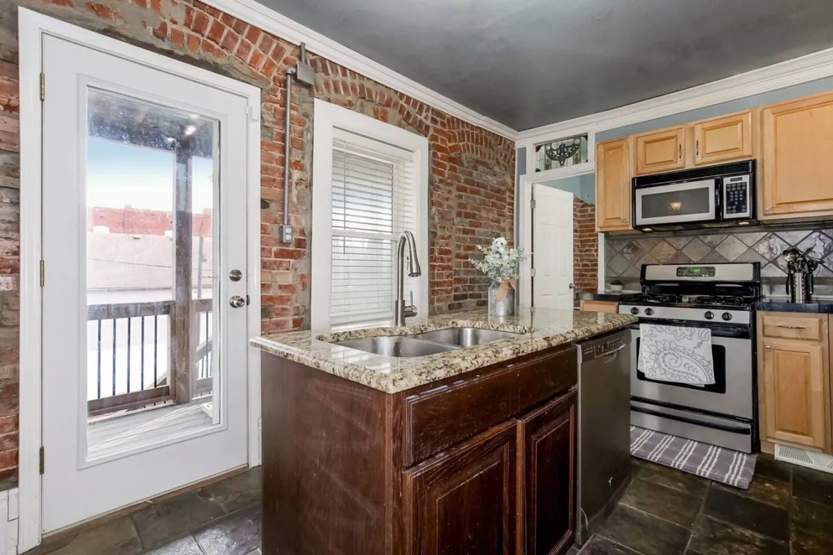 A modern kitchen boasting a granite countertop, stainless steel appliances, and exposed brick walls. The space is cozy yet functional, with enough room for families or groups to cook together.