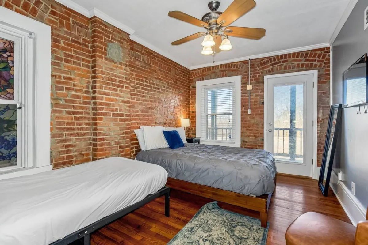 A serene bedroom featuring two comfortable beds with soft linens, an exposed brick wall, stained glass window, and double doors opening to a balcony. The space is airy and inviting, with natural light streaming in and a ceiling fan overhead for comfort.