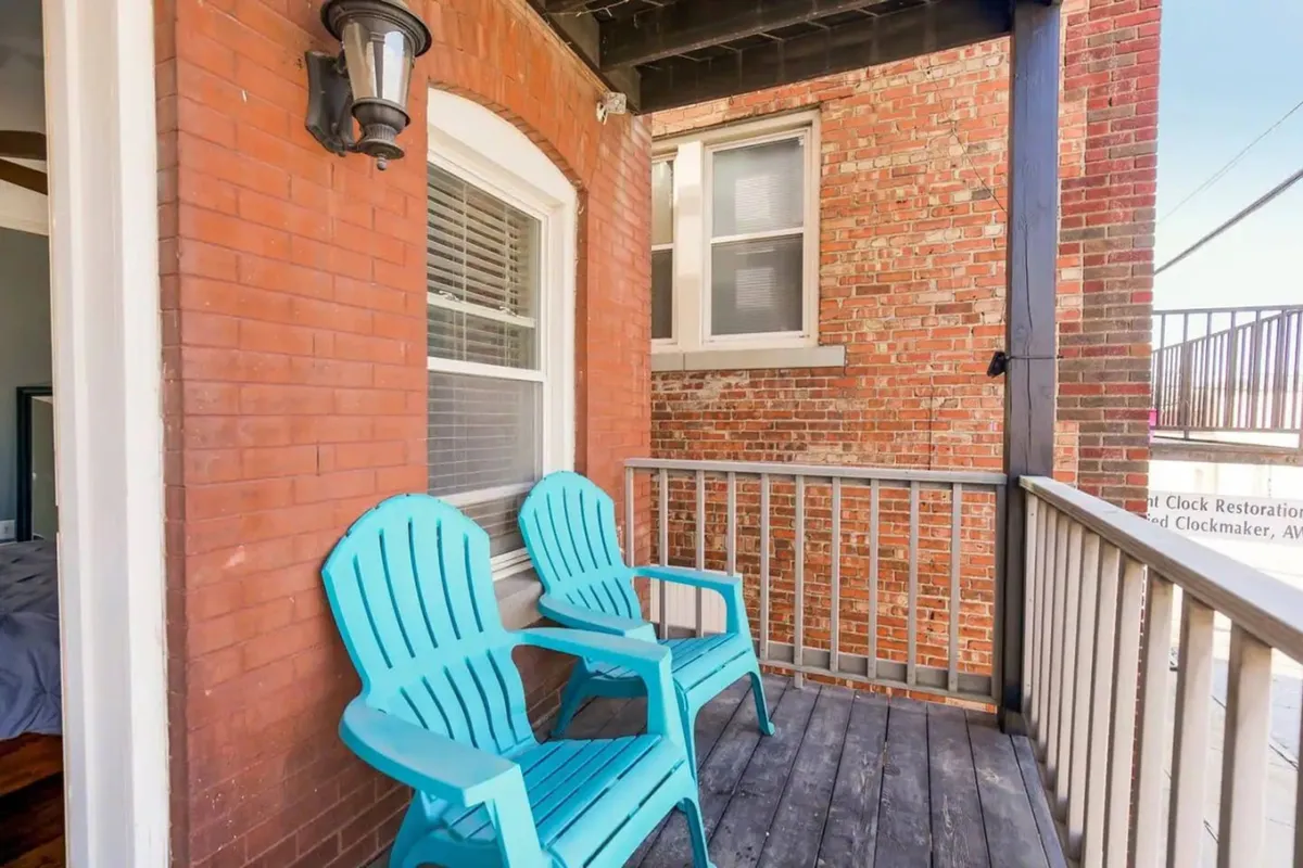 A comfortable exterior seating area with blue Adirondack chairs set against a brick wall and wooden railing, suggesting a casual spot to gather before heading out for the night. The space is ideal for relaxing, chatting with friends, or enjoying a coffee as the city wakes up.