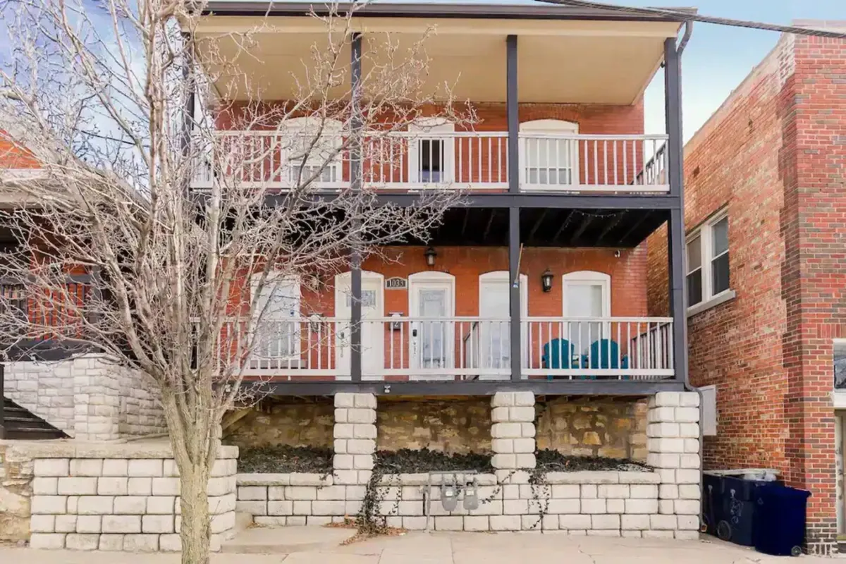 An inviting exterior balcony scene with two blue chairs, white railings, and a view of the brick building facade. The setup suggests a welcoming spot to relax, socialize, or enjoy a sunset moment.