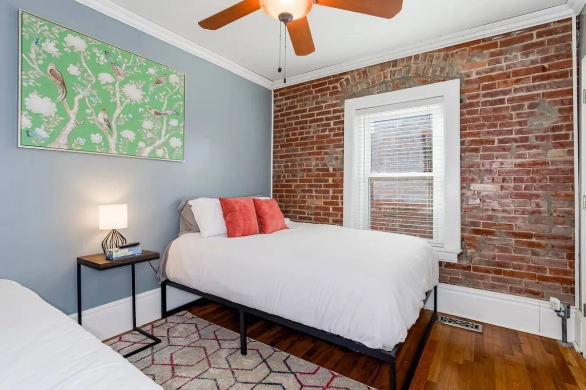 A stylish bedroom with a brick wall backdrop, an eye-catching art piece, a neatly made bed with plush bedding, and a nightstand. The space is bathed in natural light, creating a warm and inviting atmosphere.