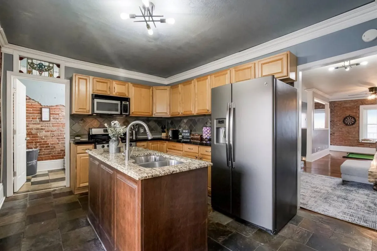 A welcoming kitchen scene with a central kitchen island, granite countertops, stainless steel appliances, exposed brick walls, and rich cabinetry. The island provides a natural gathering spot, ideal for entertaining or shared family meals.