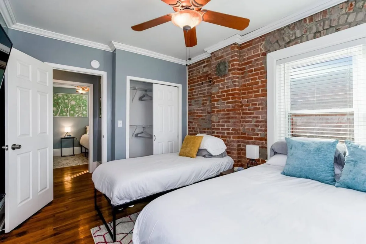 A cozy bedroom with an exposed brick wall, two beds dressed with decorative pillows, a ceiling fan, and natural light entering through windows. The room exudes relaxation and homeliness, with ample space for settling in.