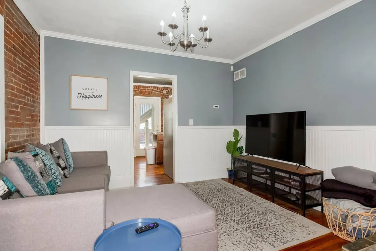 A cozy, well-lit living room featuring a plush sofa, television, striking chandelier, and wall art. The setup suggests a comfortable area for relaxing, watching TV, or enjoying conversations, with ample seating and inviting decor.