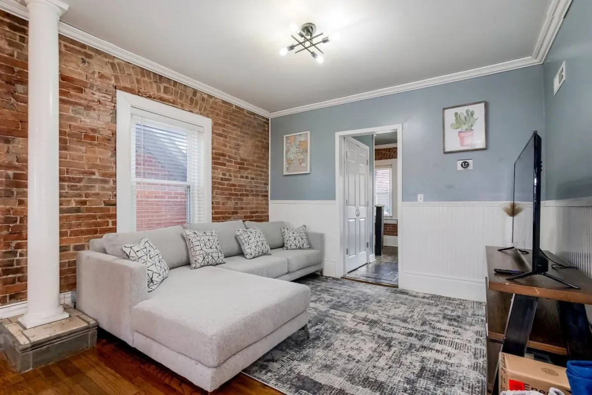 A welcoming living space featuring a sofa, brick wall, television, window, and artwork. The combination of seating and natural light from the window provides a cozy yet vibrant atmosphere, perfect for conversation and unwinding.