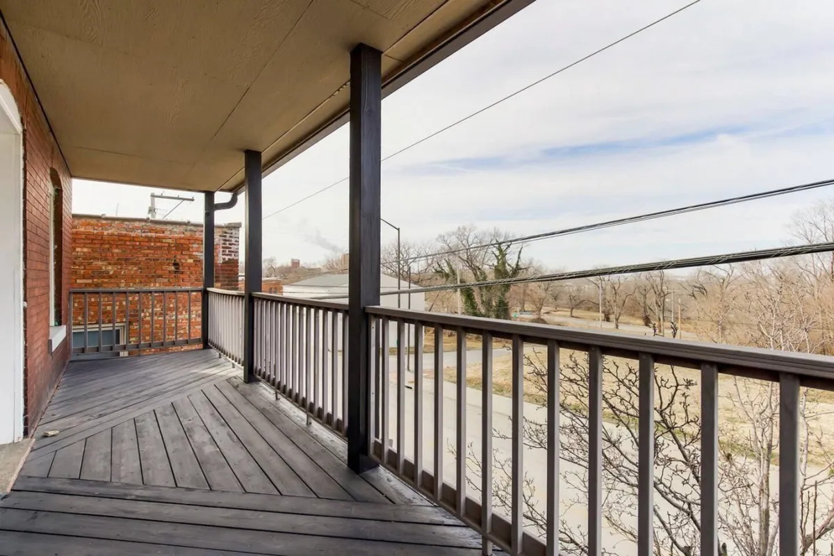 A tranquil exterior balcony space with wooden railing, surrounded by mature trees and open sky. The setting is quiet and private, offering a peaceful spot to relax, socialize, or enjoy the neighborhood views.
