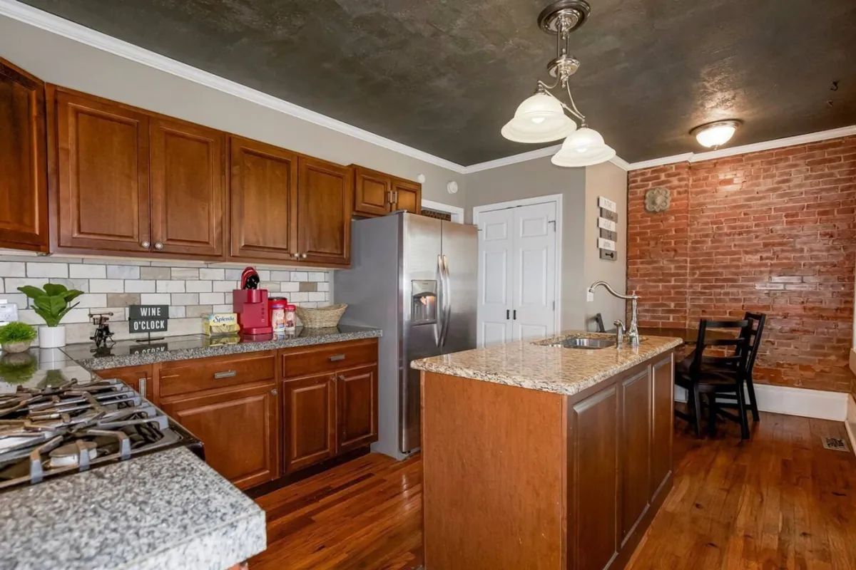 A spacious kitchen featuring a granite countertop, stainless steel appliances, exposed brick walls, and warm wooden cabinetry. The area is clearly set up for cooking and entertaining, with plenty of counter space for meal prep and space for gatherings.