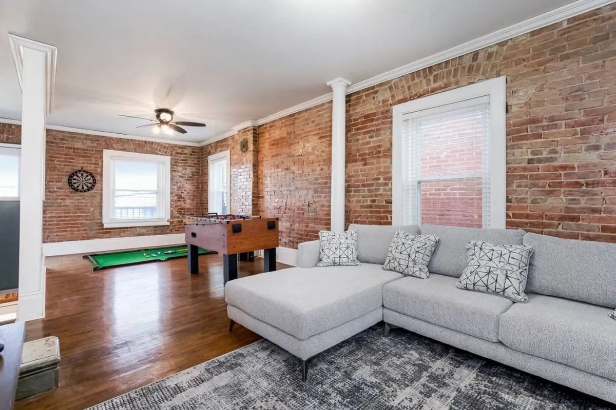 The image showcases a spacious, light-filled living area with exposed brick walls, a stylish modern sectional sofa, and large windows. The room features a foosball table and dartboard, suggesting a fun, social atmosphere perfect for gatherings. The overall look is both playful and comfortable, inviting guests to relax, play games, and enjoy quality time together.