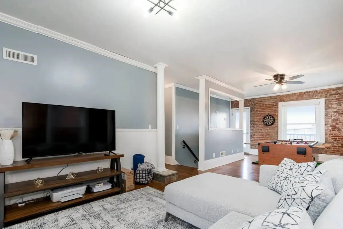 A spacious living area with a plush sofa, large TV, brick wall, and a foosball table. The room is designed for both relaxation and entertainment, offering options for watching movies or enjoying a friendly game night.