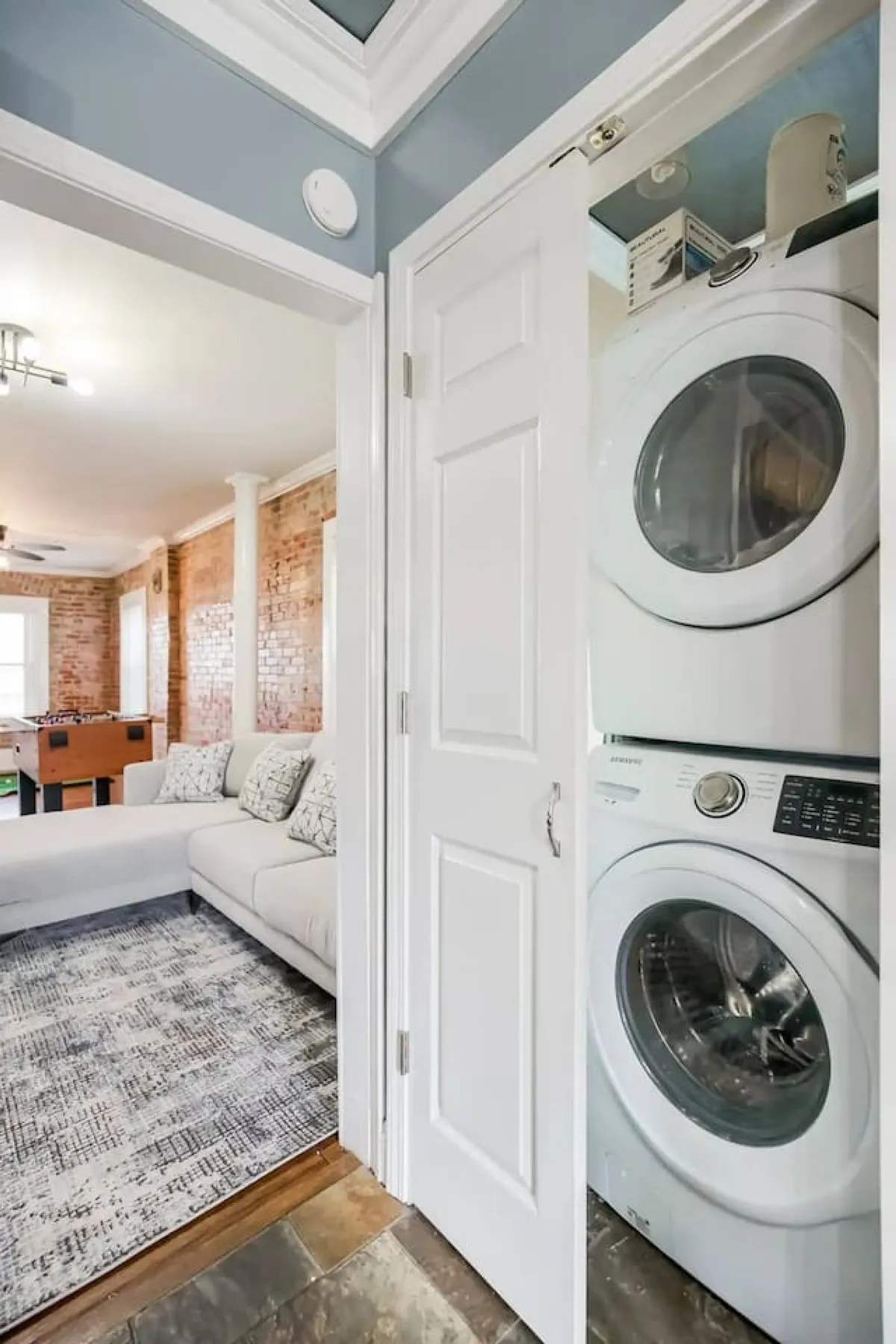 A versatile laundry area with a washer and dryer set, a couch, brick wall, area rug, and foosball table. The image emphasizes the ease of doing laundry in a cozy, multi-purpose space that encourages relaxation between chores.